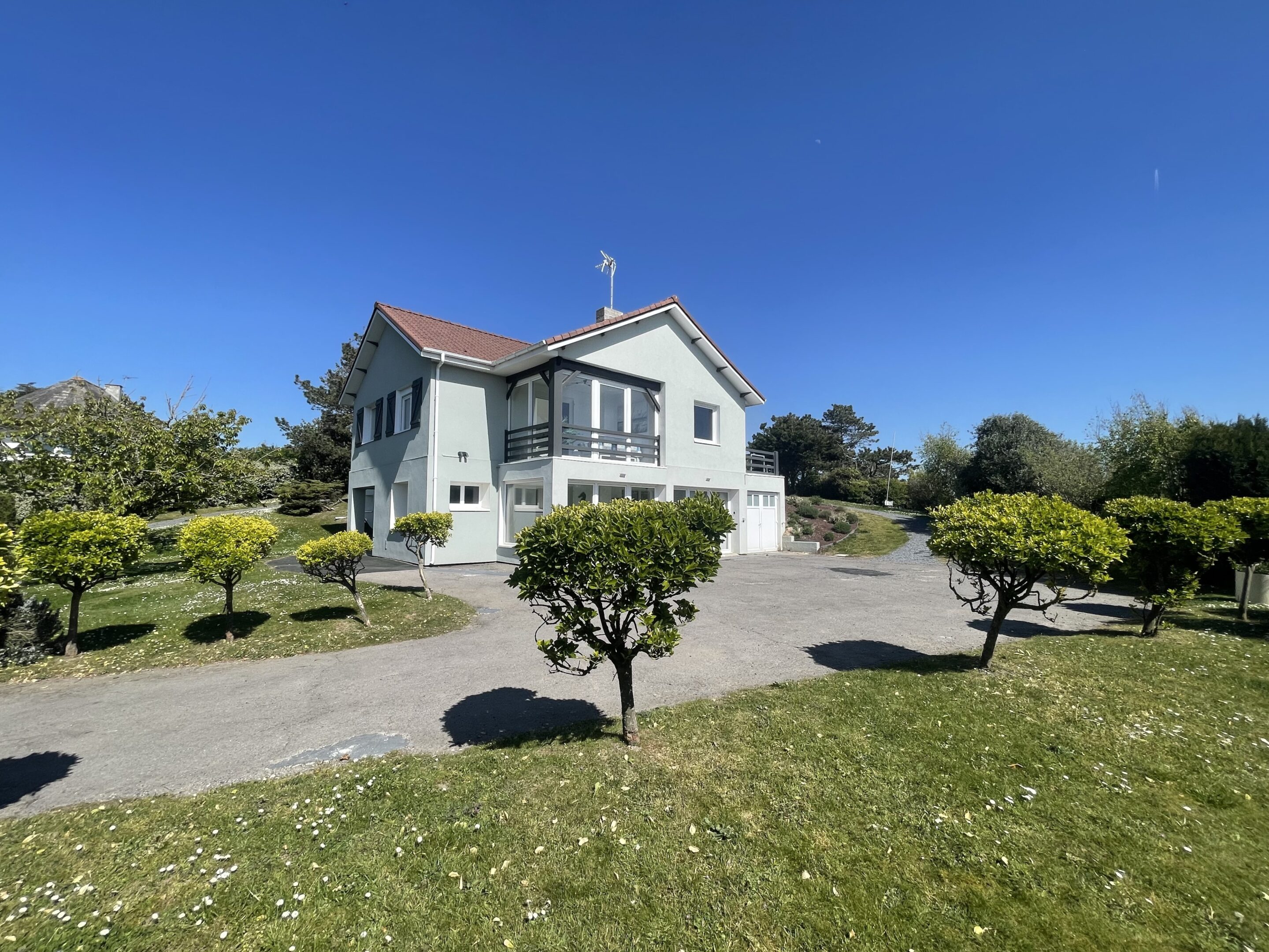 Two-story light-gray house with a red-tiled roof and a small balcony, set in a manicured garden under a clear blue sky, with an antenna on the roof.
