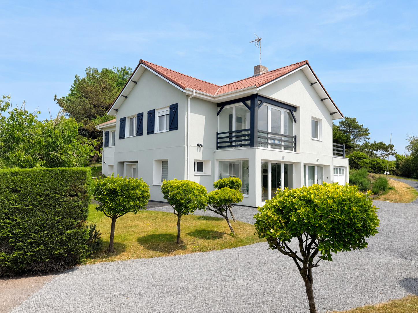 White two-story house with a red-tiled roof, blue shutters, and a glass-enclosed balcony, set among greenery and a gravel driveway.